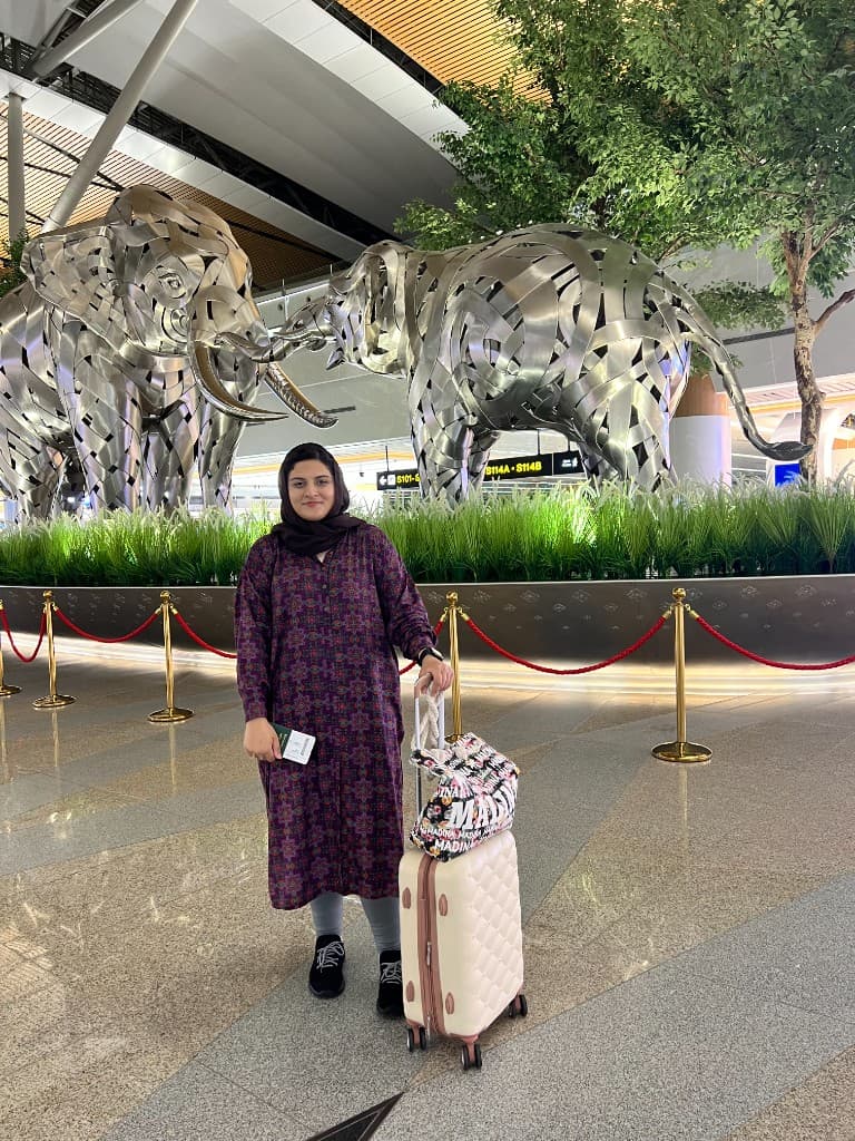 Kainat Kamal with passport and luggage in an airport terminal, representing global movement and Her Kadam’s gateway for women.